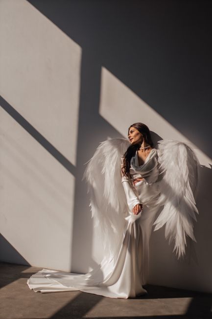 A woman in a white dress with wings, bathed in dramatic sunlight, creating angelic shadows indoors.