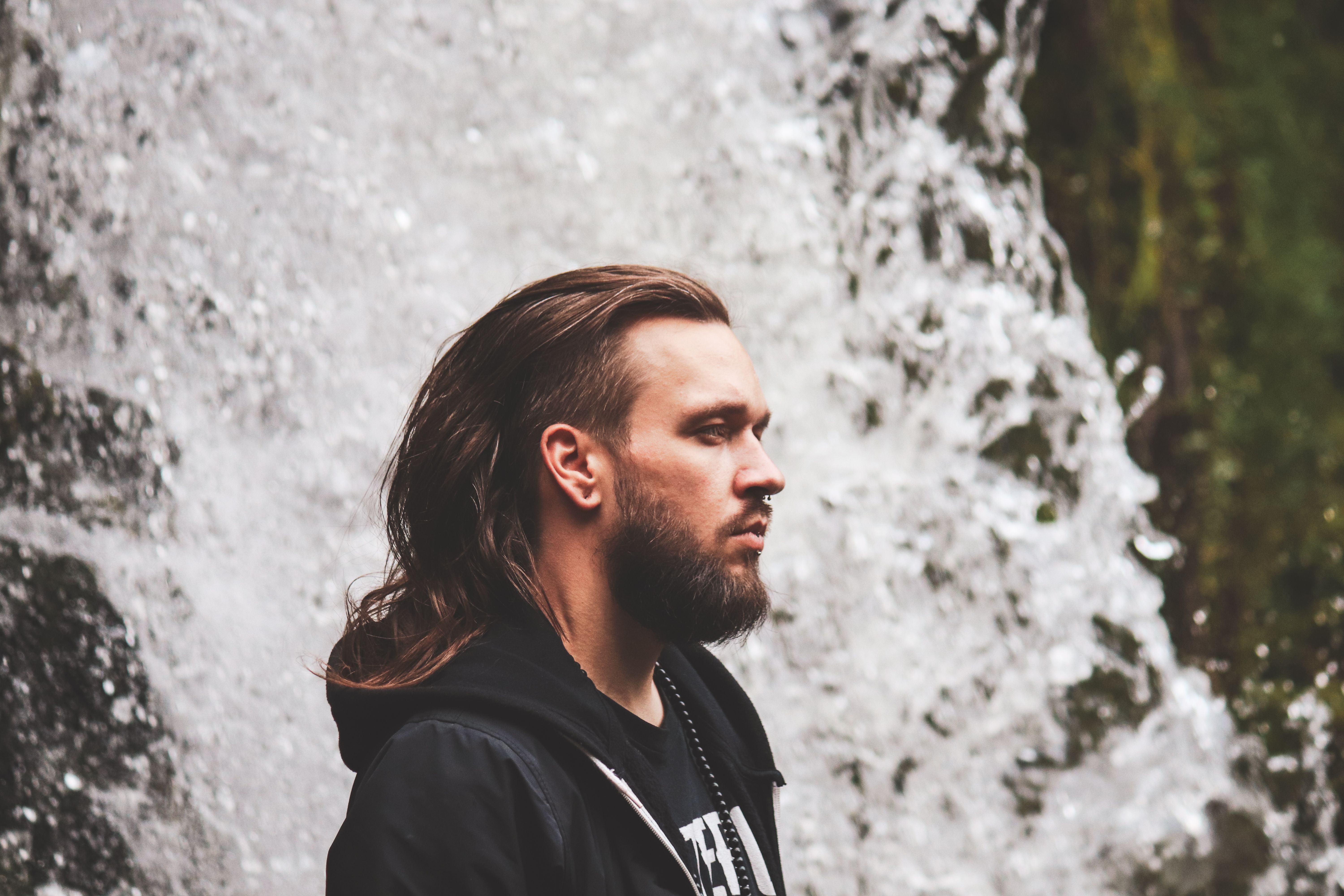 Profile of a bearded man in a hoodie standing near a waterfall.
