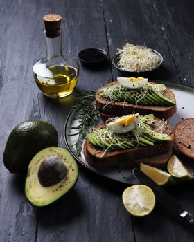 Avocado toast topped with egg and sprouts served with lemon and olive oil on a wooden table.