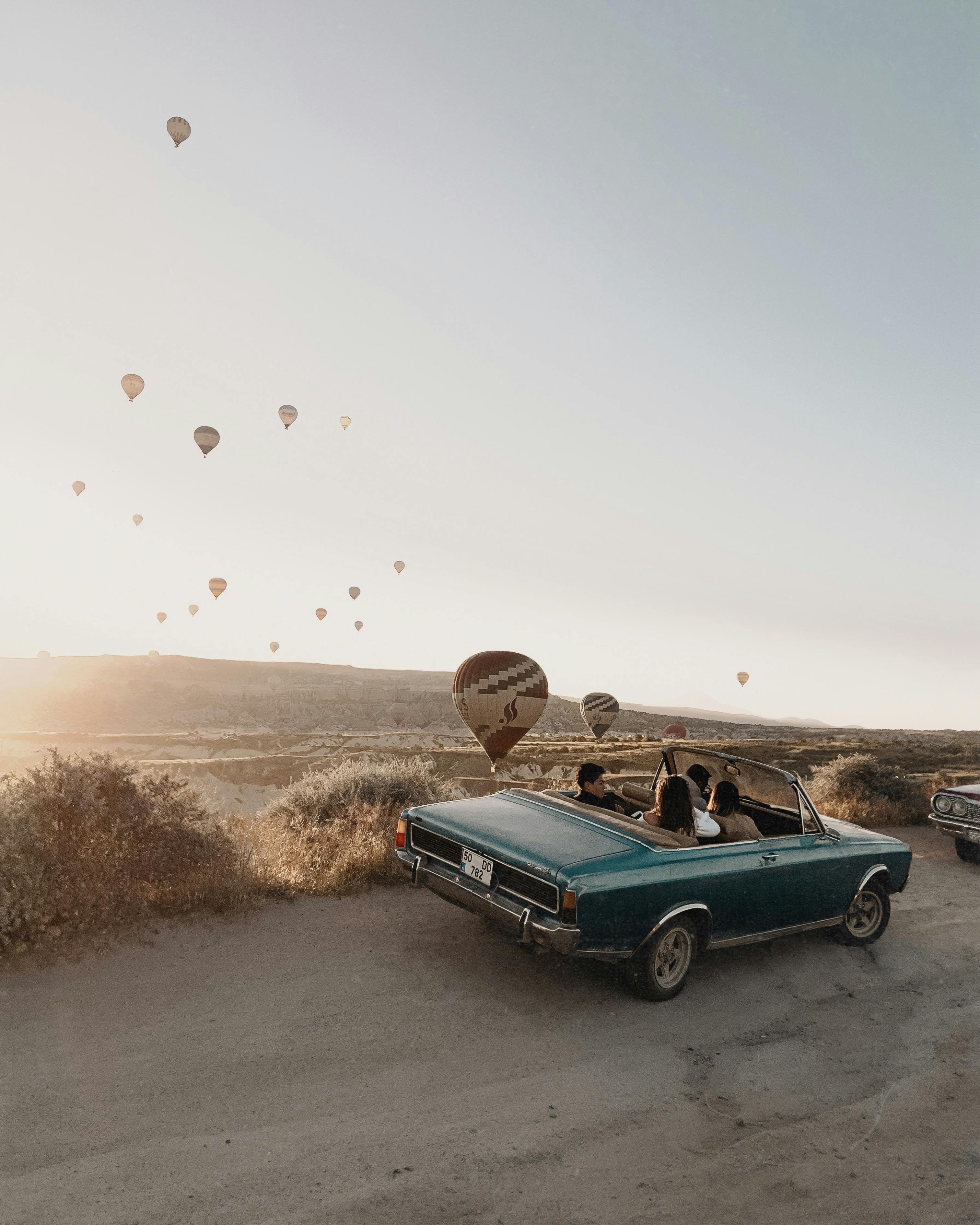 A vintage convertible car with passengers enjoying a scenic drive in Cappadocia amidst hot air balloons at sunrise.