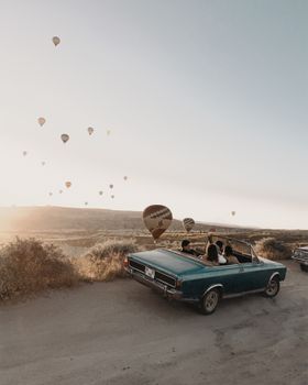 A vintage convertible car with passengers enjoying a scenic drive in Cappadocia amidst hot air balloons at sunrise.