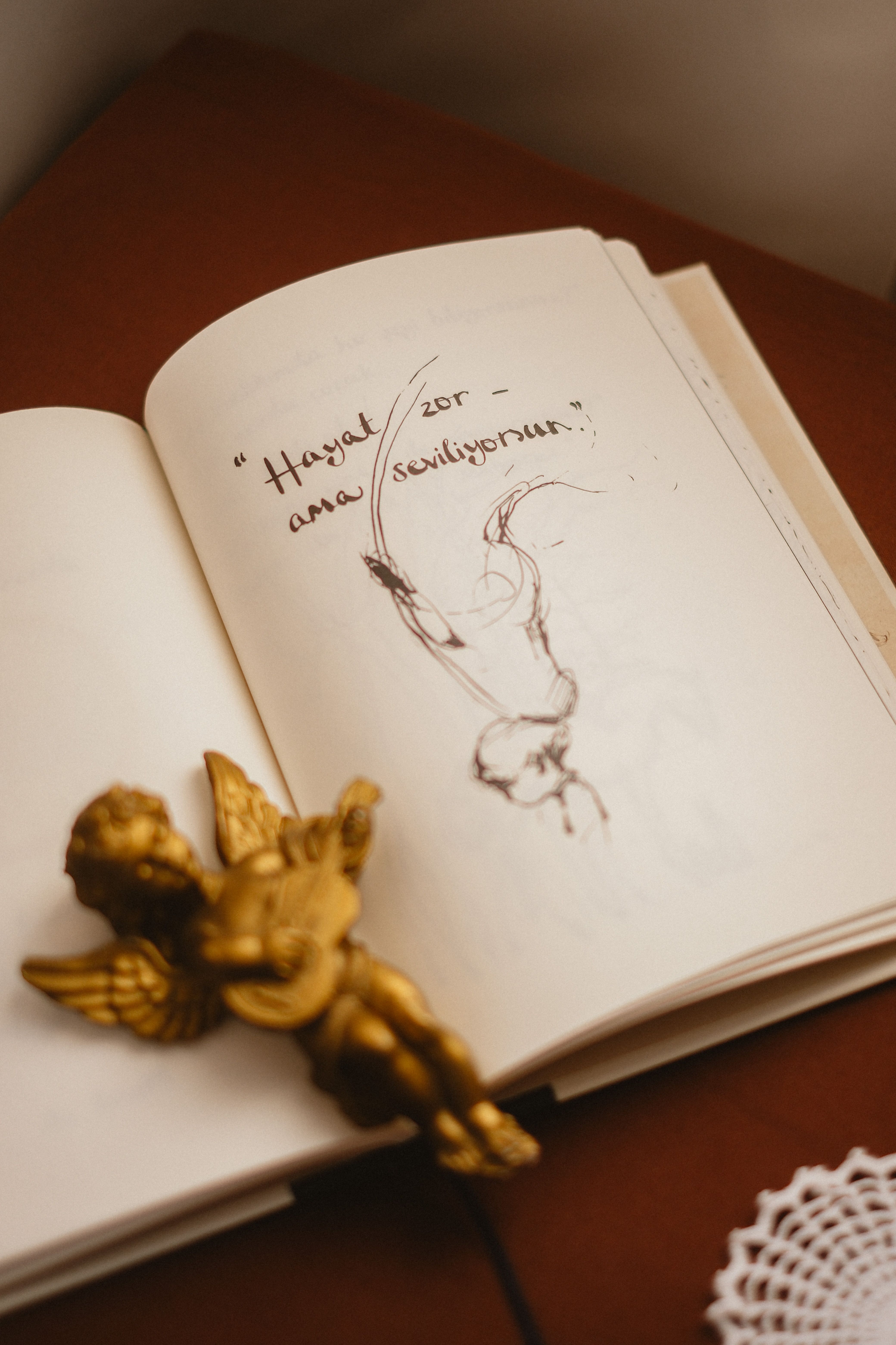Still life of an open book with a sketched figure, gold angel figurine, and elegant quote.