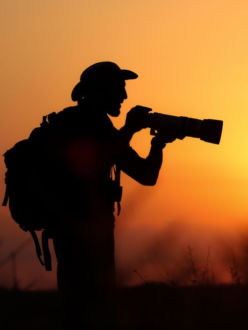 Free Silhouette of a photographer with hat and camera in hand against a vivid sunset sky. Stock Photo