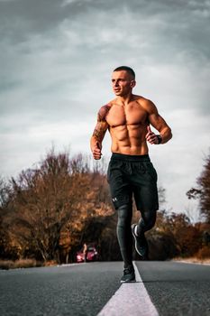 A shirtless man jogging on a road surrounded by autumn foliage, showcasing fitness and outdoor exercise.