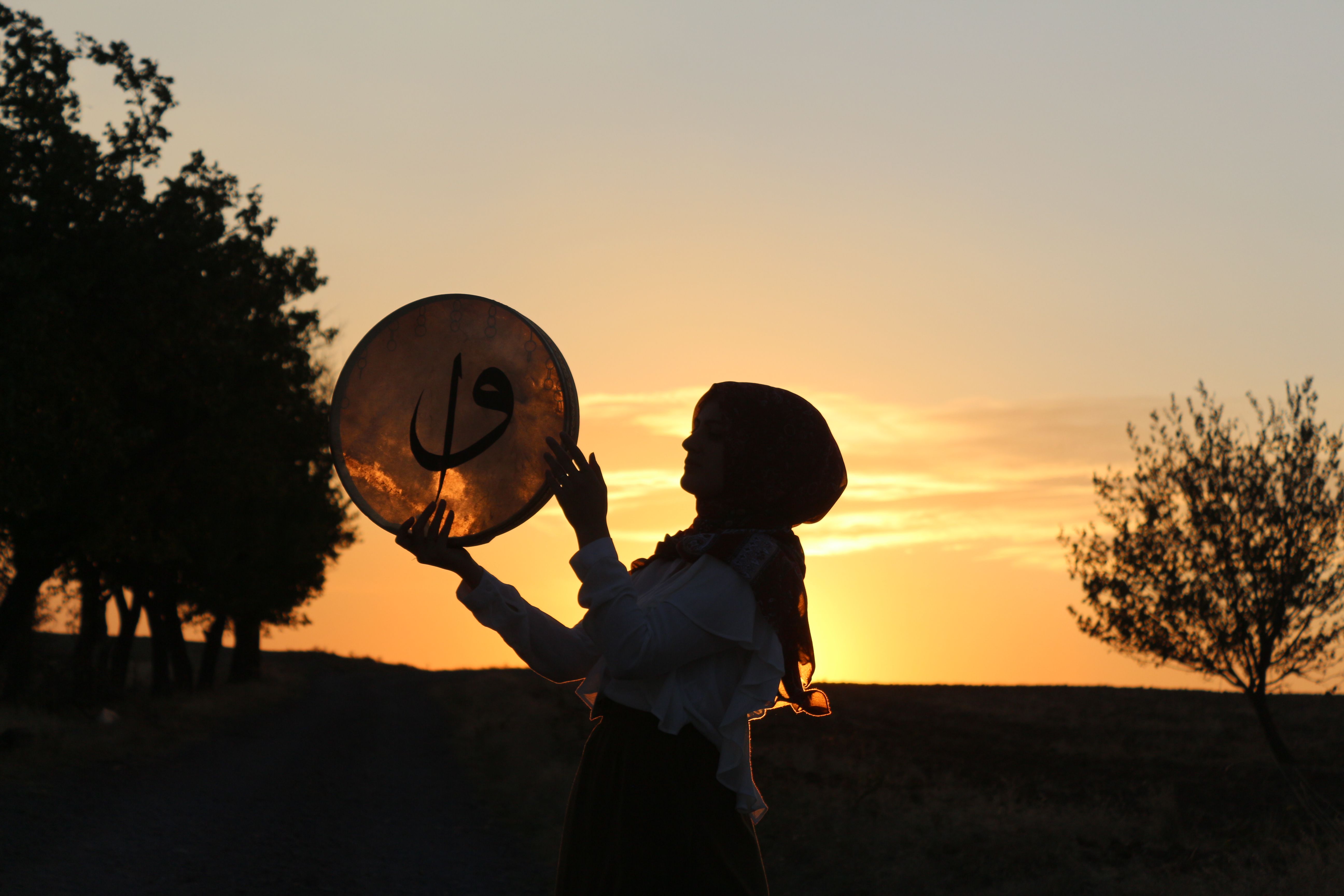 Silhouette of woman in hijab holding a traditional drum against a stunning sunset backdrop.