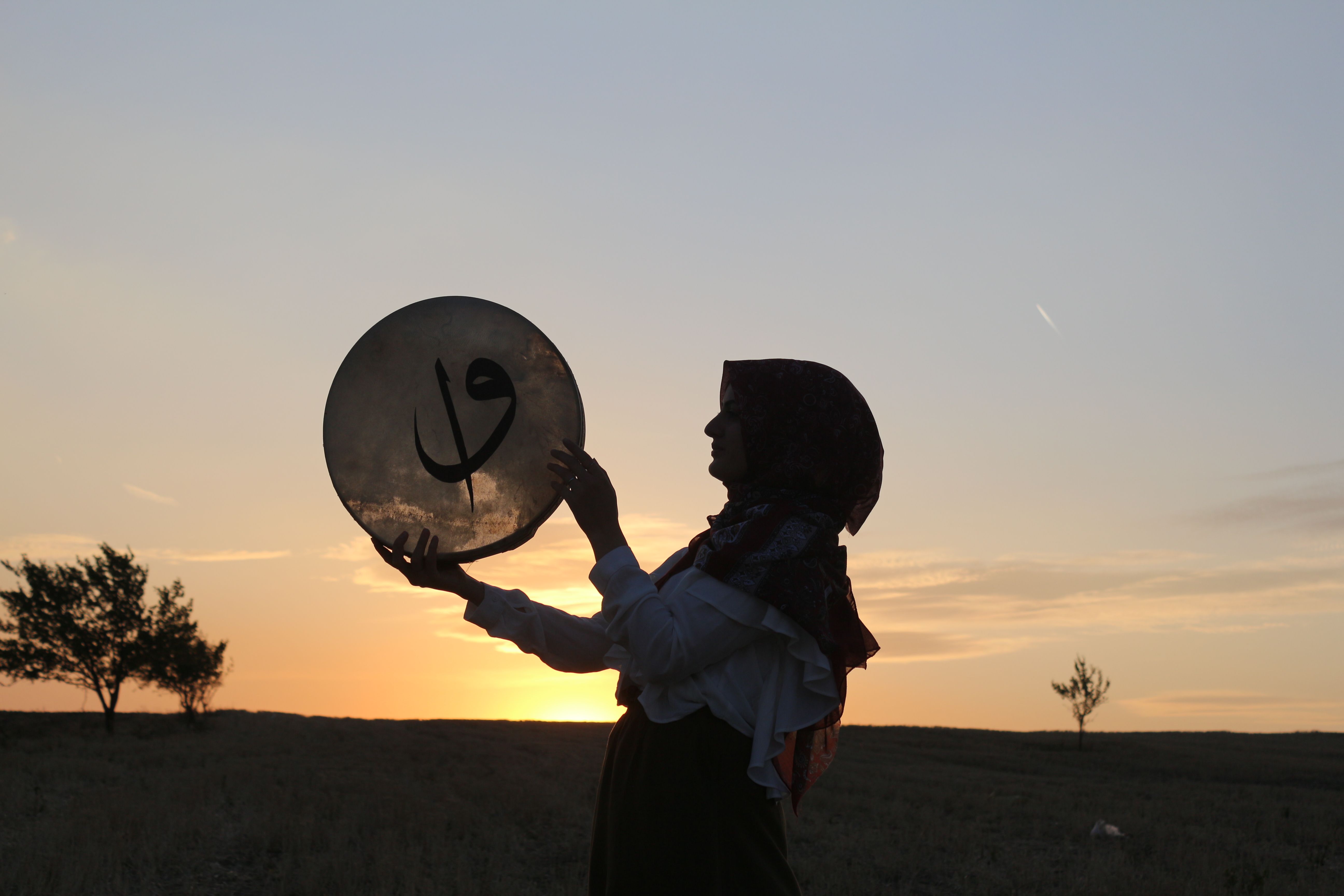 Silhouette of a woman holding a drum against a vibrant sunset in a rural landscape. Perfect for cultural or musical themes.
