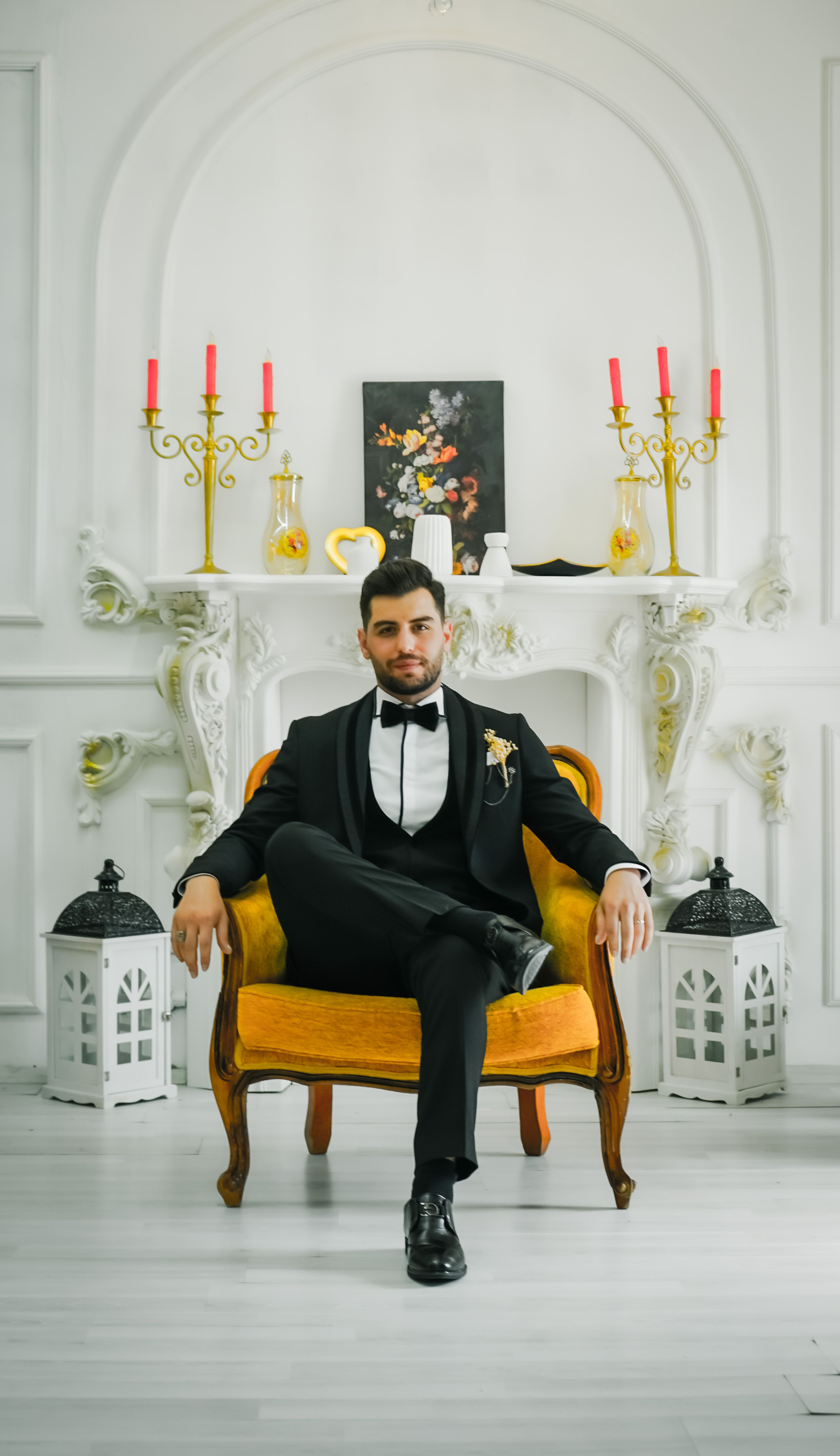 Un mire elegant îmbrăcat stă pe un fotoliu într-un decor interior clasic și luxos, cu elemente decorative ornate.