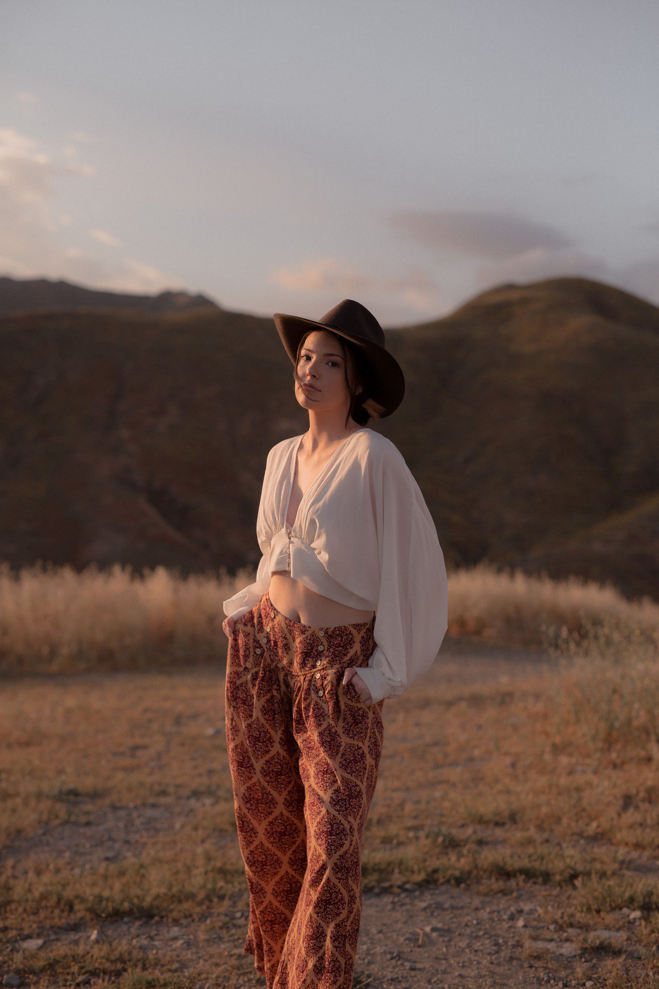 Uma mulher fashion vestindo um chapéu apreciando o pôr do sol em um cenário ao ar livre.