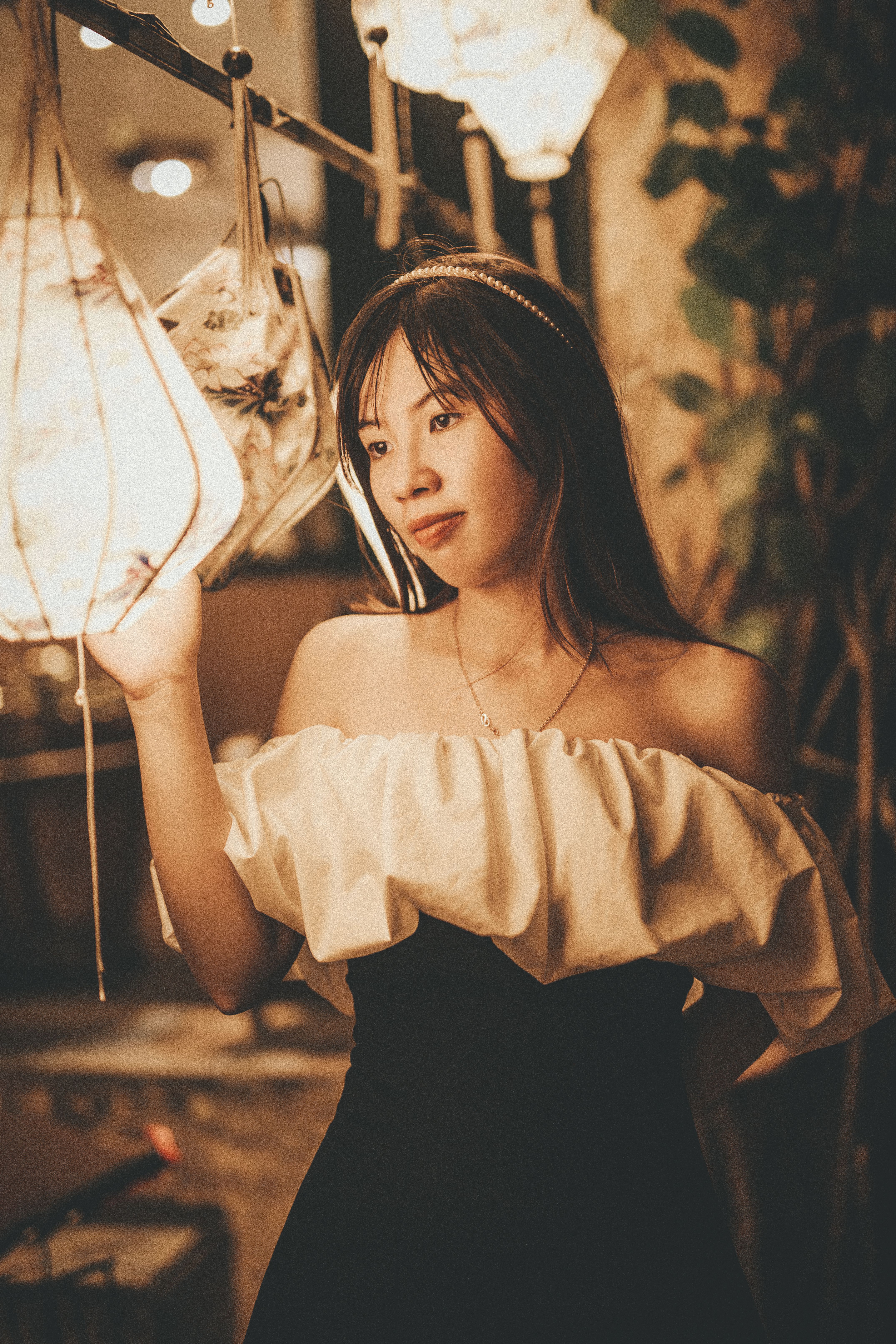 Chic portrait of an Asian woman standing indoors touching a glowing lamp, evoking warmth and elegance.