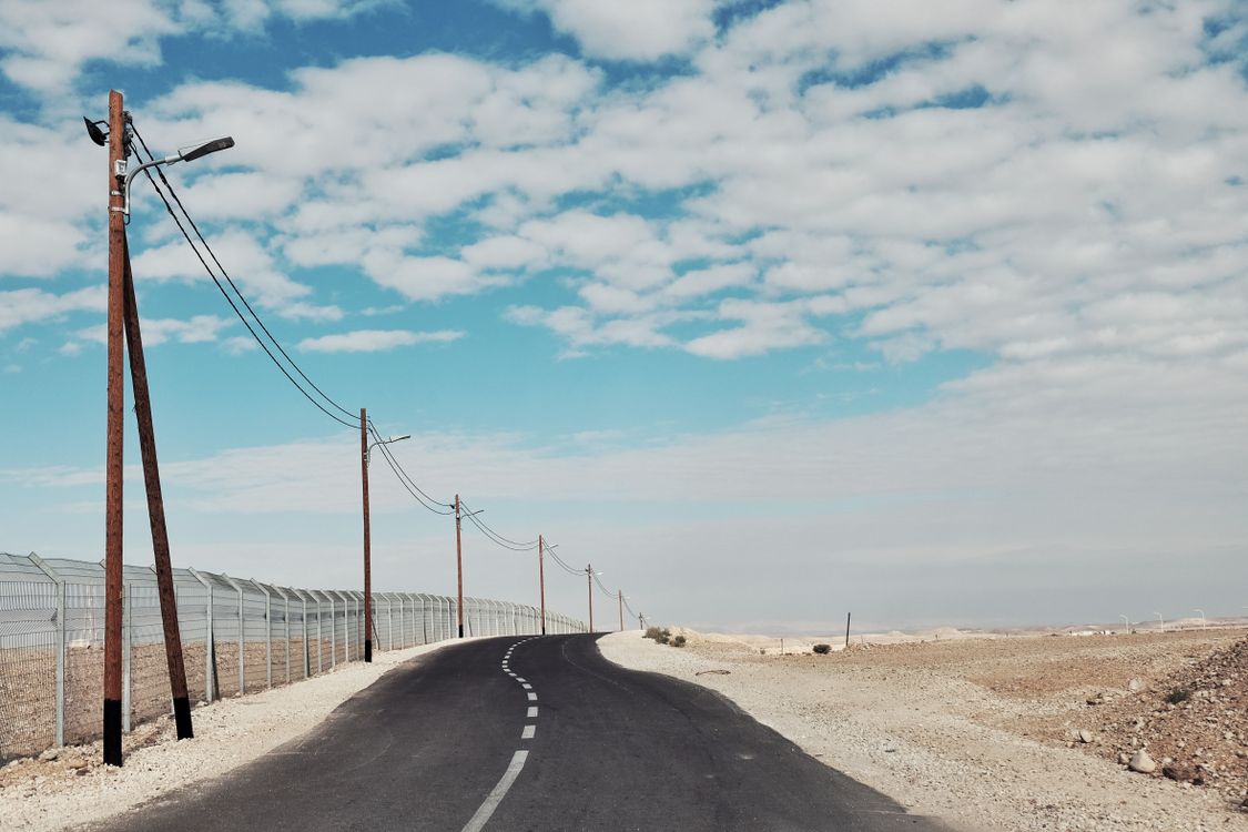Desert road with utility poles and a fence line under a partly cloudy sky