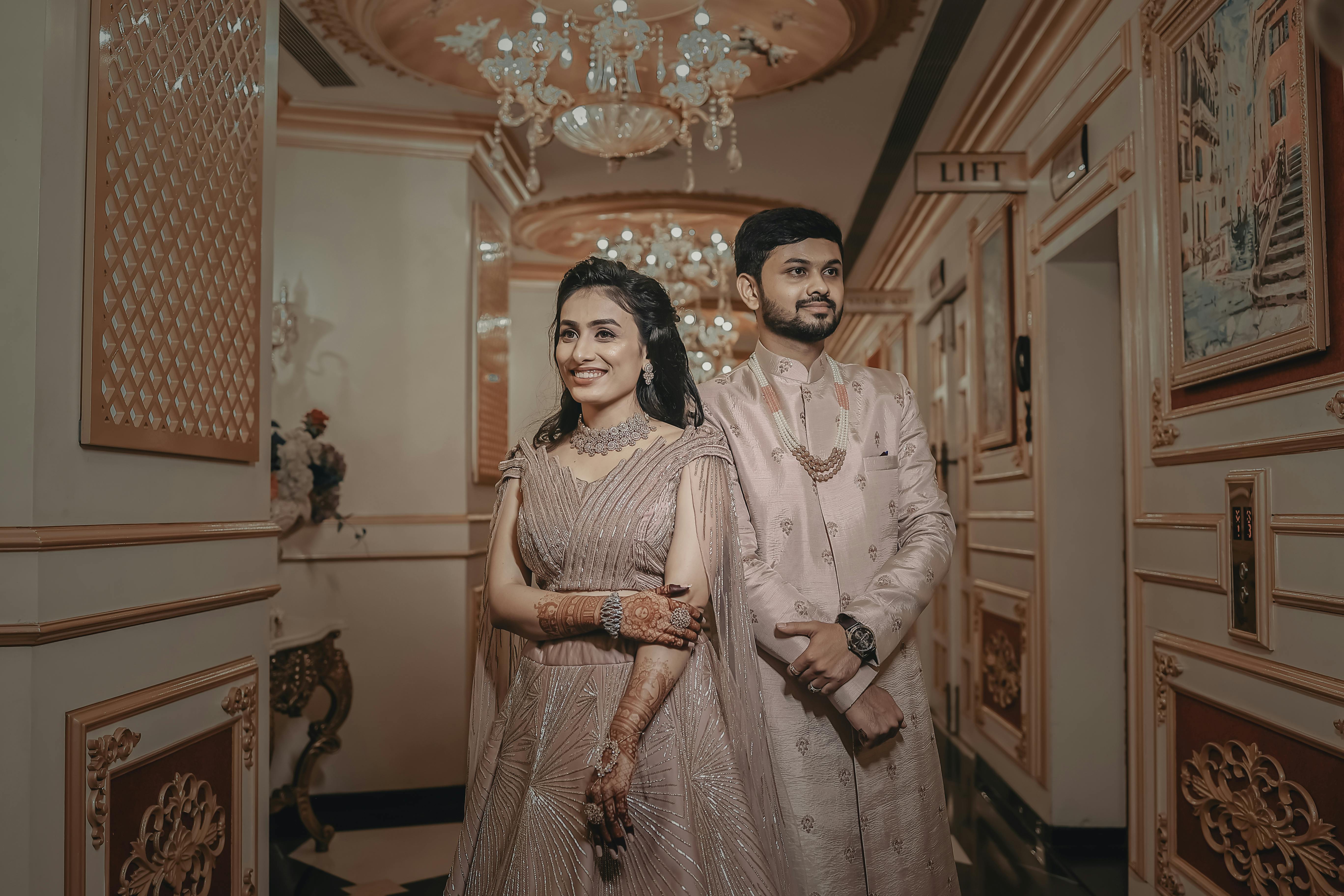 A smiling couple in elegant Indian attire stands indoors with luxurious decor.