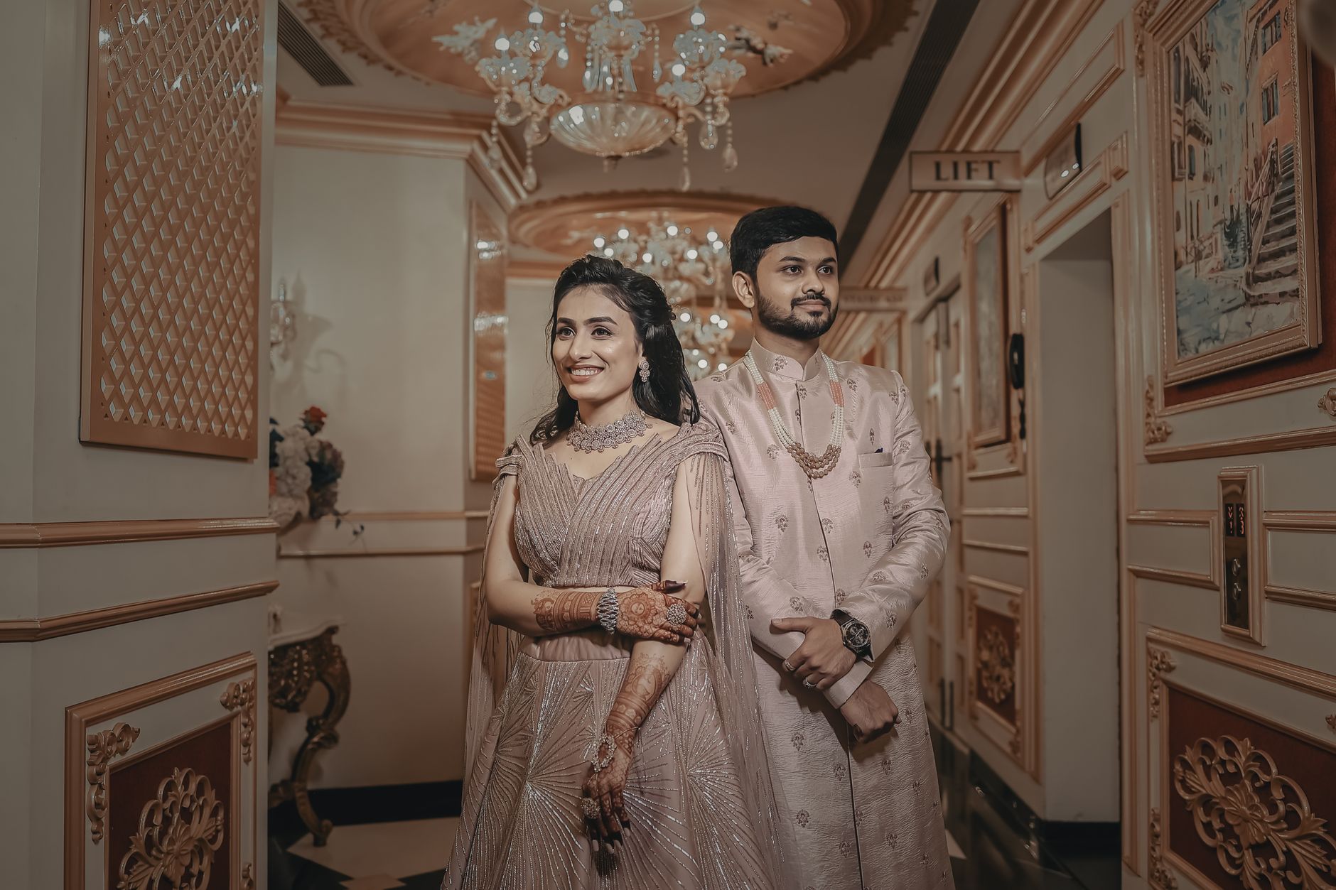 A smiling couple in elegant Indian attire stands indoors with luxurious decor.