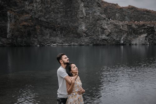 Free A couple embraces by a serene lake with rocky cliffs under a cloudy sky. Stock Photo