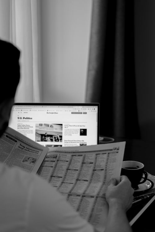 Free Black and white photo of a man reading a newspaper with a laptop in the background. Stock Photo
