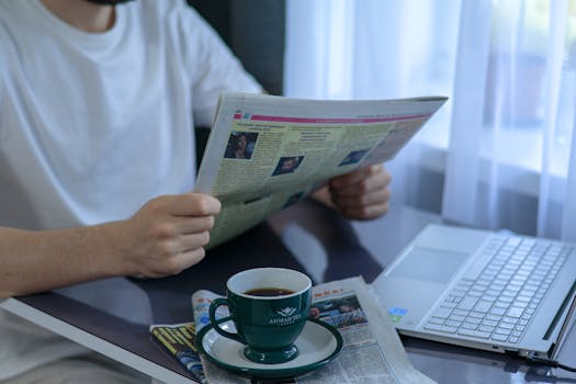 Adult enjoying coffee and newspaper at a desk with laptop. Indoors, bright daylight setting.