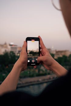 Hands holding a smartphone capturing a cityscape at sunset, with a blurred urban background.