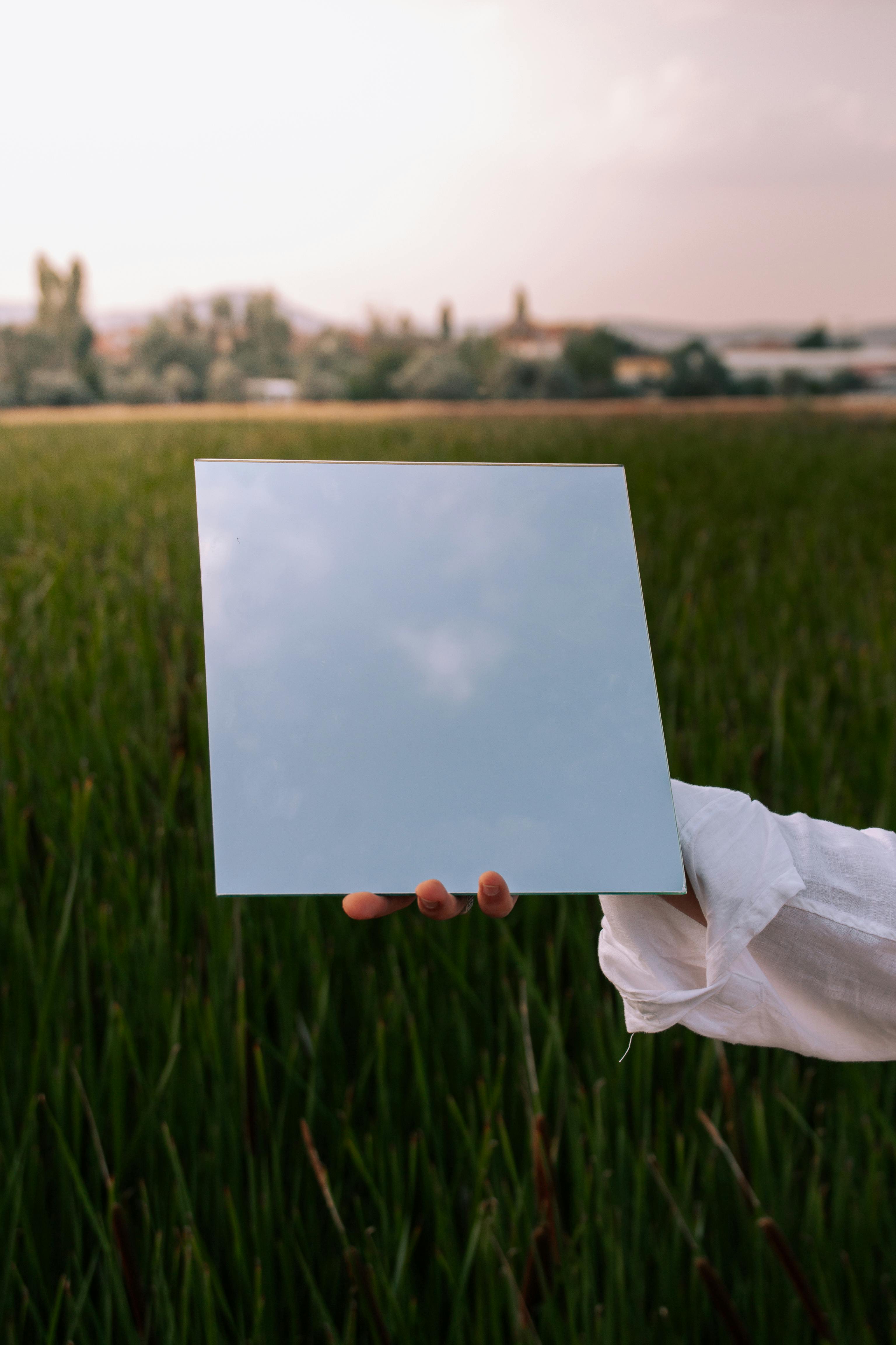 Hand holding a mirror in a rural field reflecting the sky, creating a surreal effect.