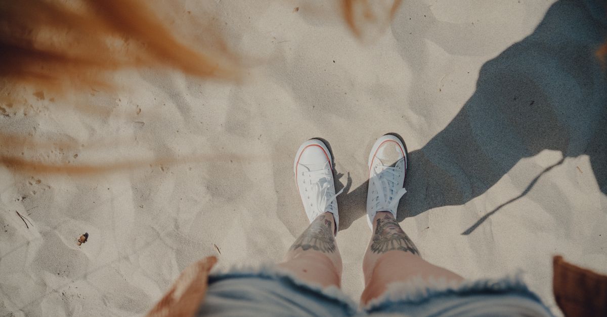 Top down view of legs in denim shorts and sneakers on sandy beach. Vibrant tattoos visible.