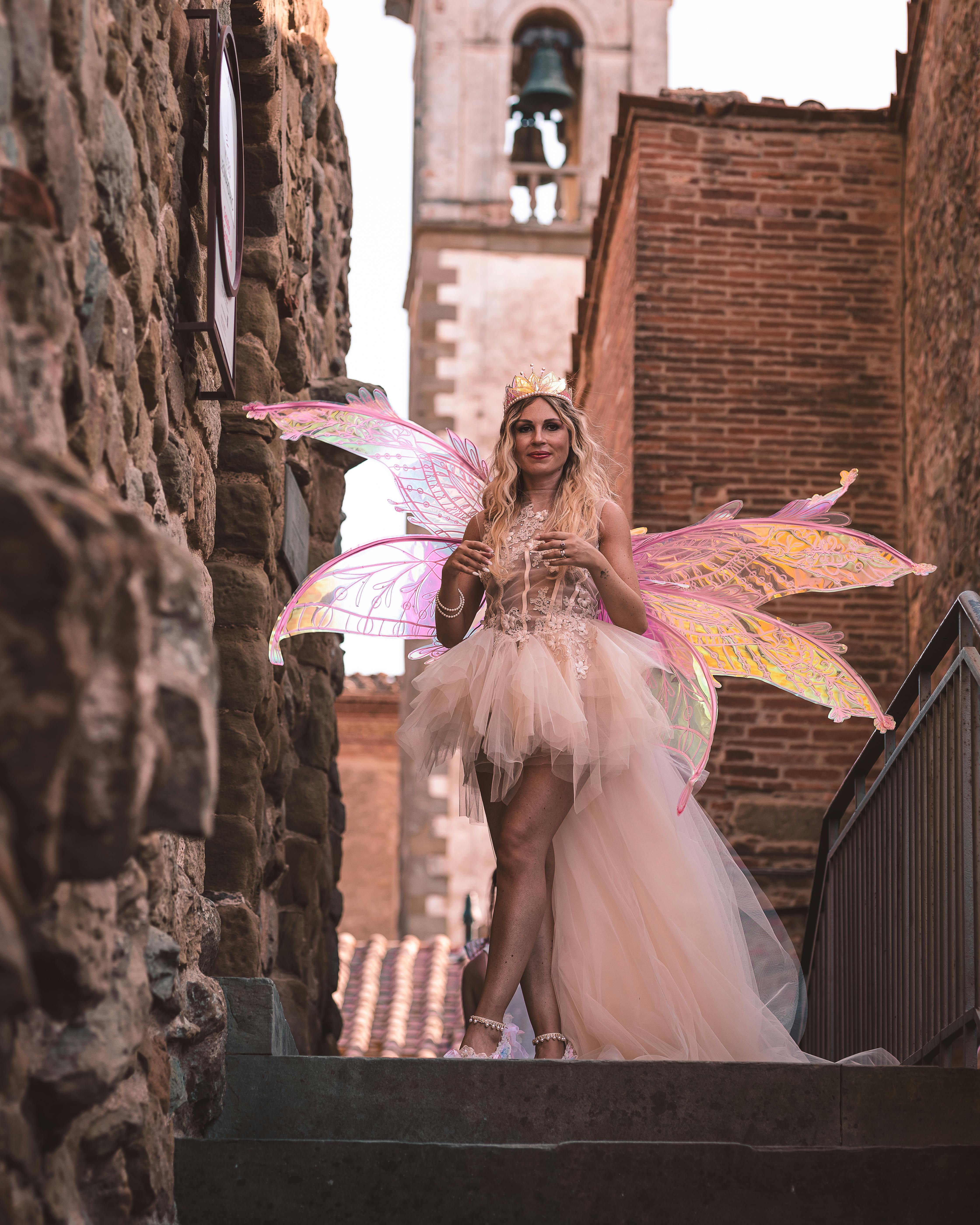 Woman in intricate fairy costume standing in urban alleyway with bell tower in the background.