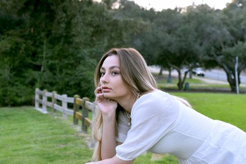 Free Portrait of a young woman with long hair leaning on a fence in a lush, green park. Stock Photo