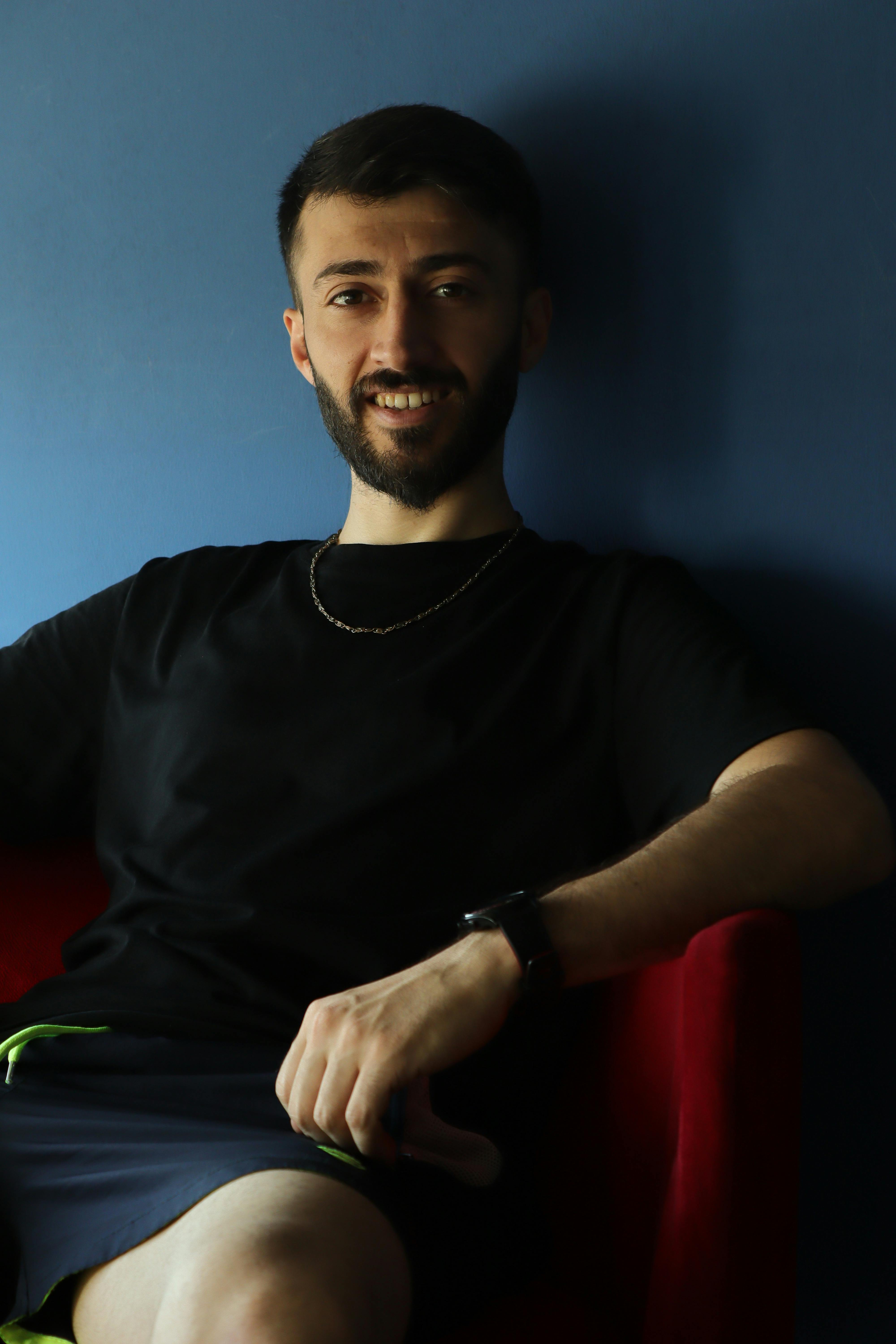 A young man sitting comfortably indoors, wearing a casual outfit with a warm smile.