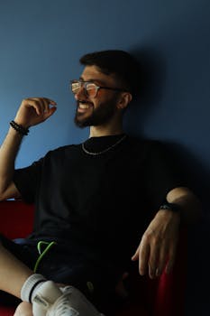 Bearded man wearing glasses smiles while sitting against a blue wall.