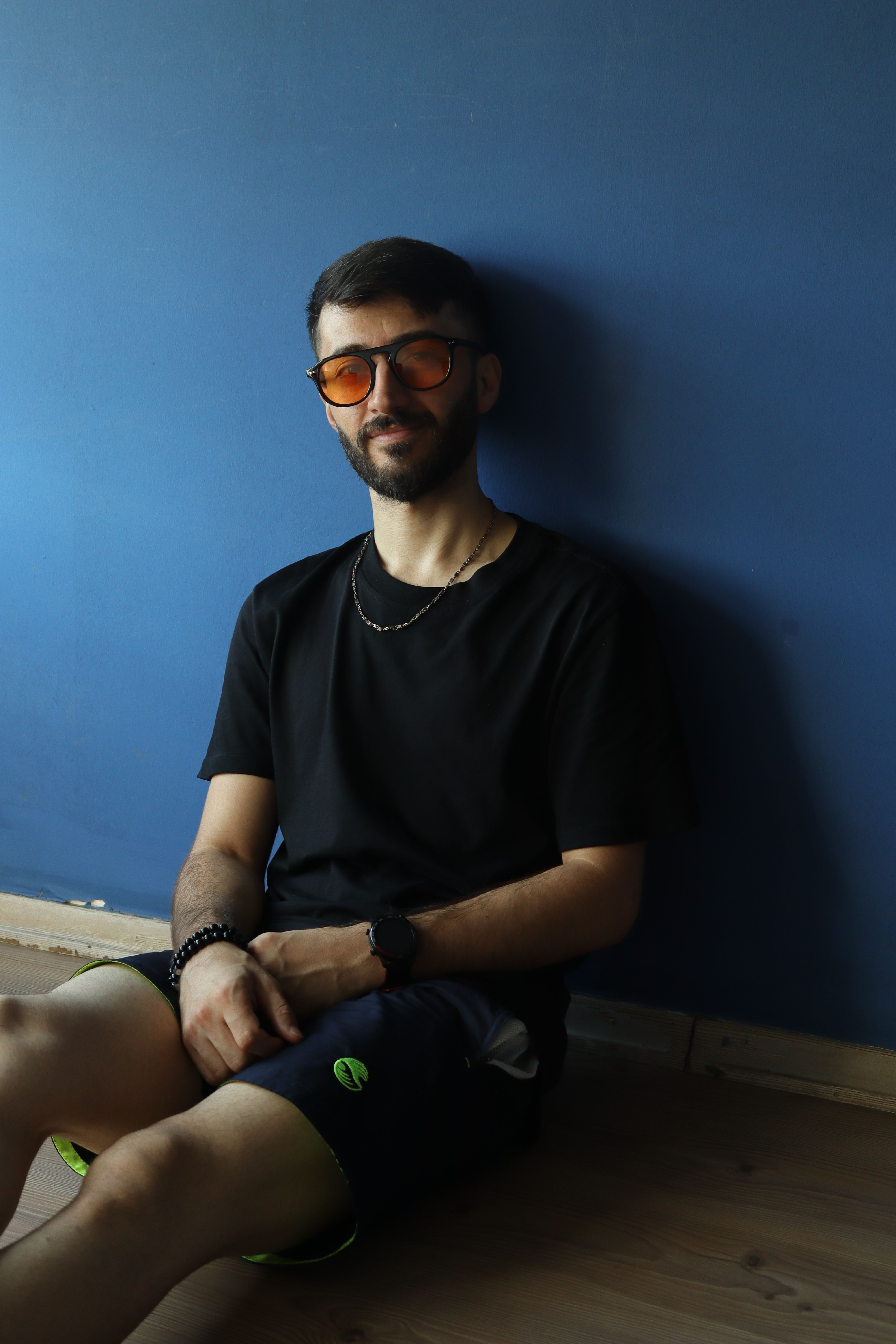 Stylish man in orange sunglasses sitting casually indoors against a blue wall.