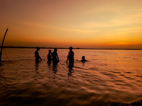 Silhouetted teenagers swimming together in a lake during a vibrant sunset, creating a joyful summer scene.