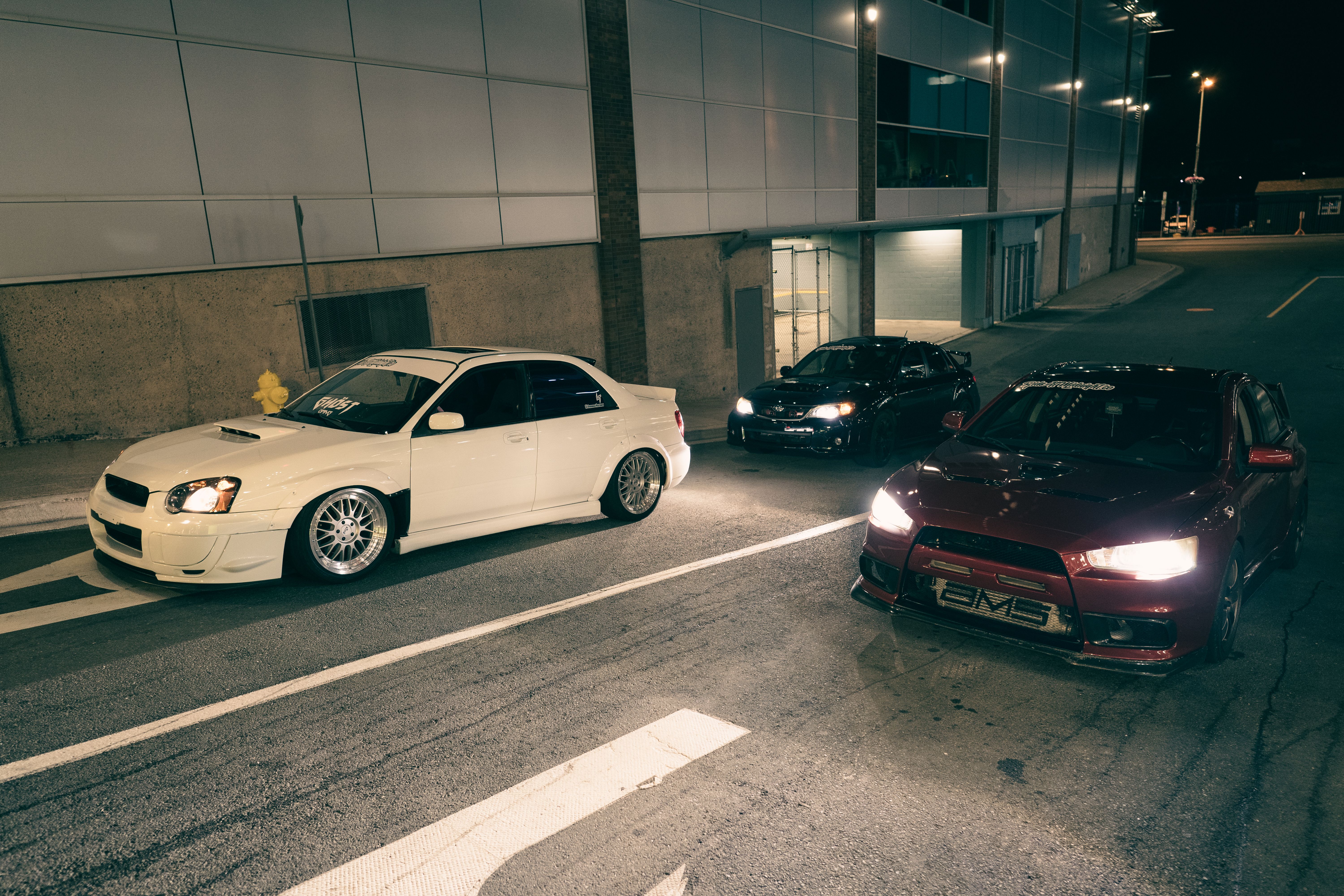 Three modified cars parked on an urban street at night, showcasing automotive style.