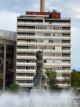 A statue stands in front of a high-rise office building with urban architectural elements on a cloudy day.