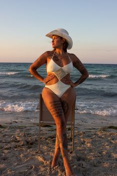 Woman poses confidently in a white swimsuit and cowboy hat during a beach sunset.