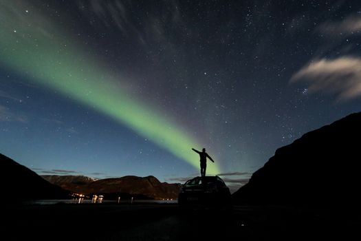 A man stands on a car, silhouetted against the stunning aurora borealis and starry night sky.