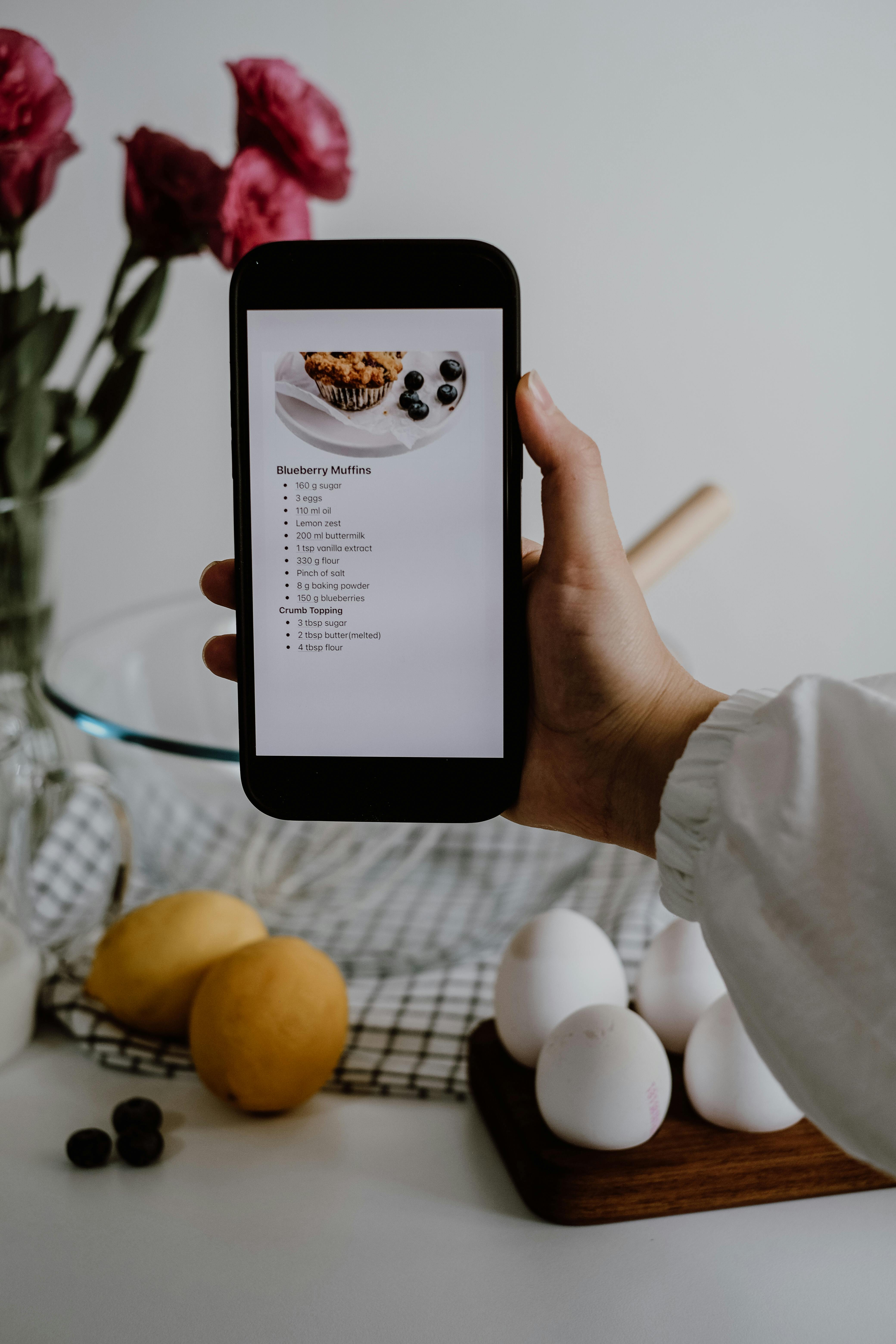 A smartphone with a blueberry muffin recipe held over kitchen ingredients.