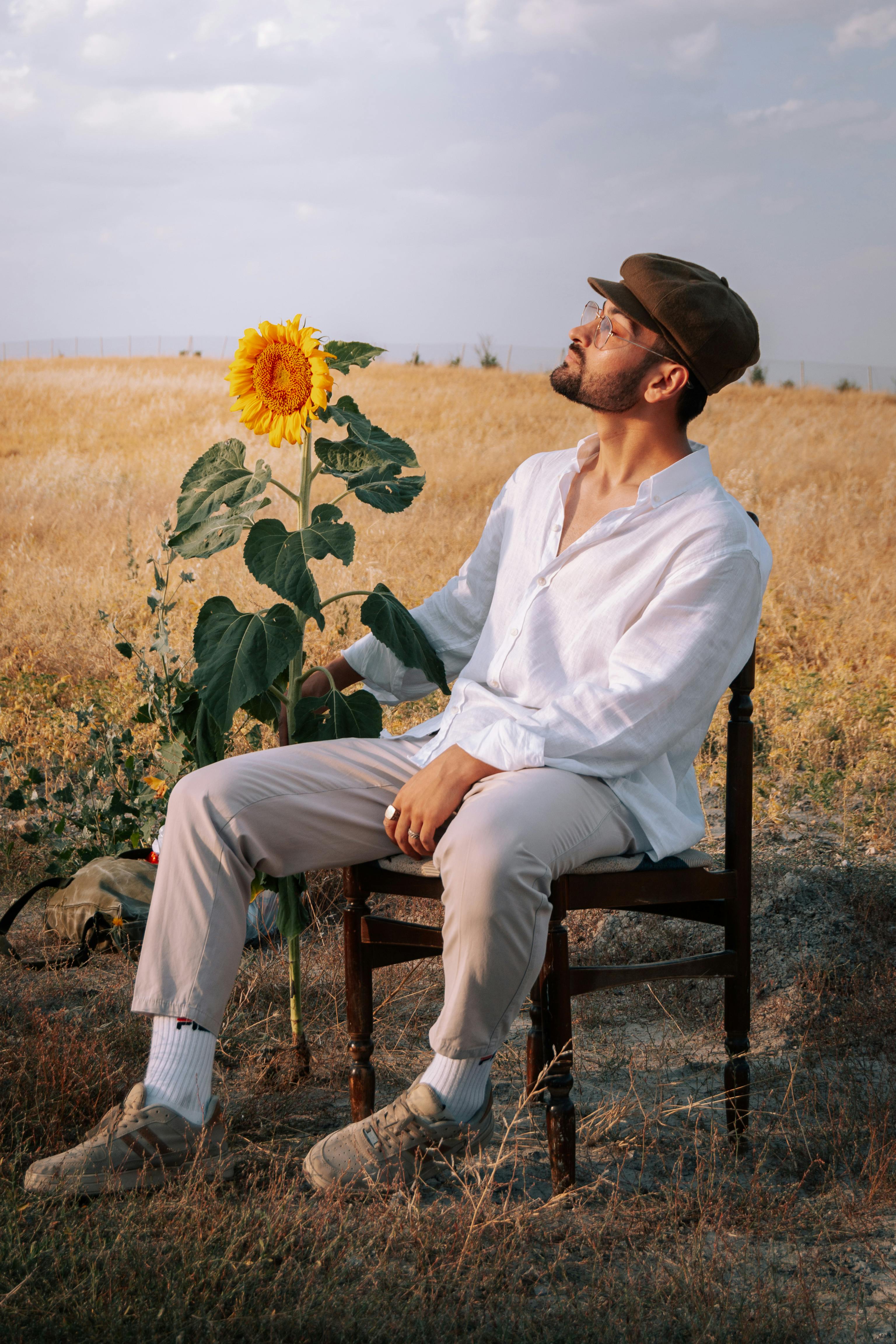 A man in a white shirt and hat sits by a sunflower in a dry field at sunset.