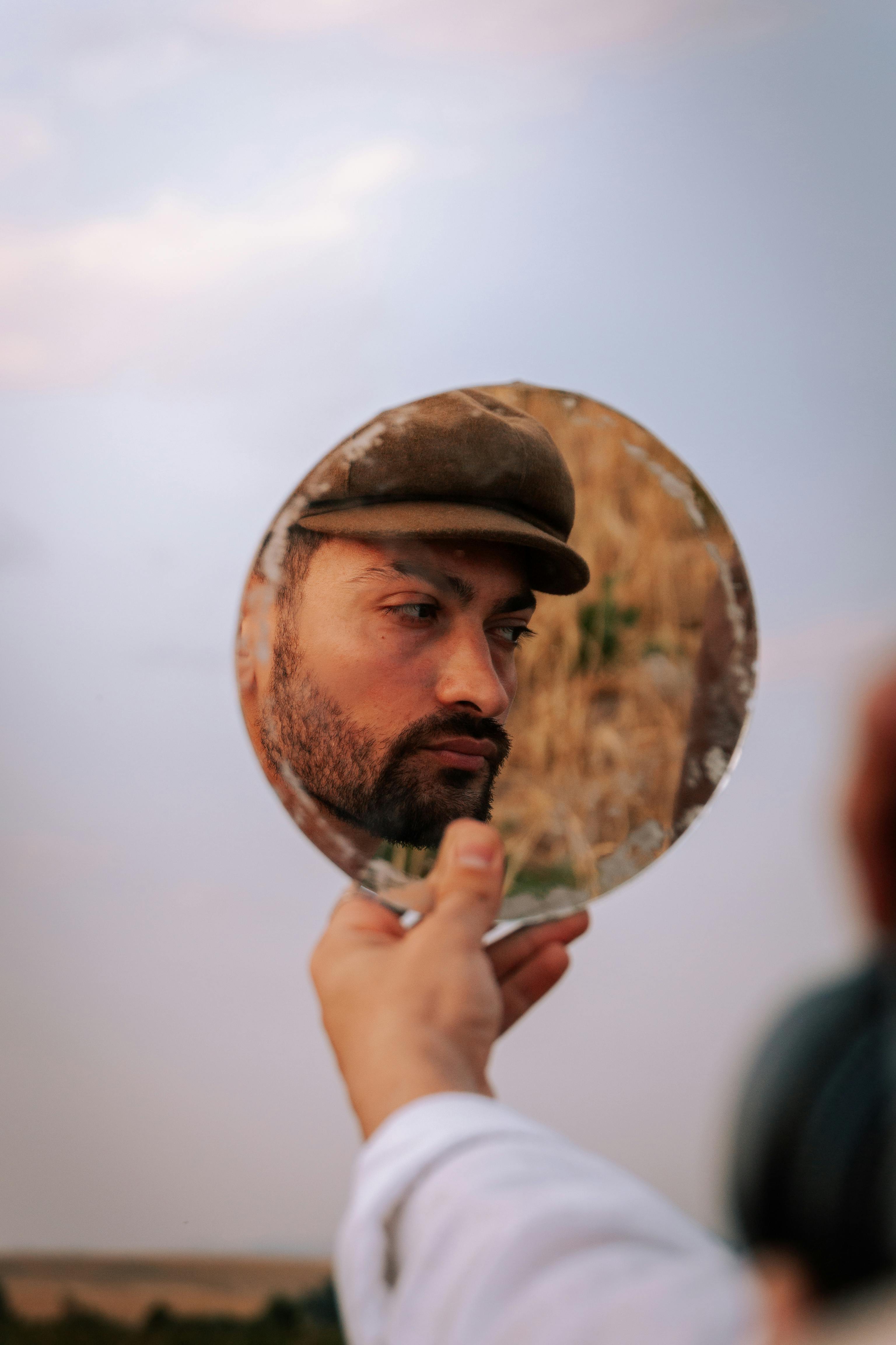 Man's reflection in a handheld mirror outdoors, thoughtful expression.