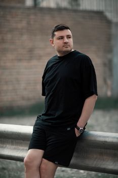 A young man in casual wear leaning against a rail in an urban setting during the day.