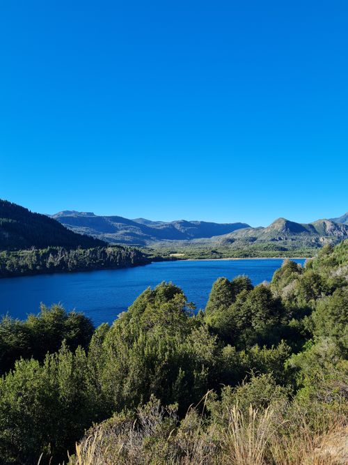 Free Breathtaking view of a serene mountain lake surrounded by lush greenery and majestic hills under a clear blue sky. Stock Photo