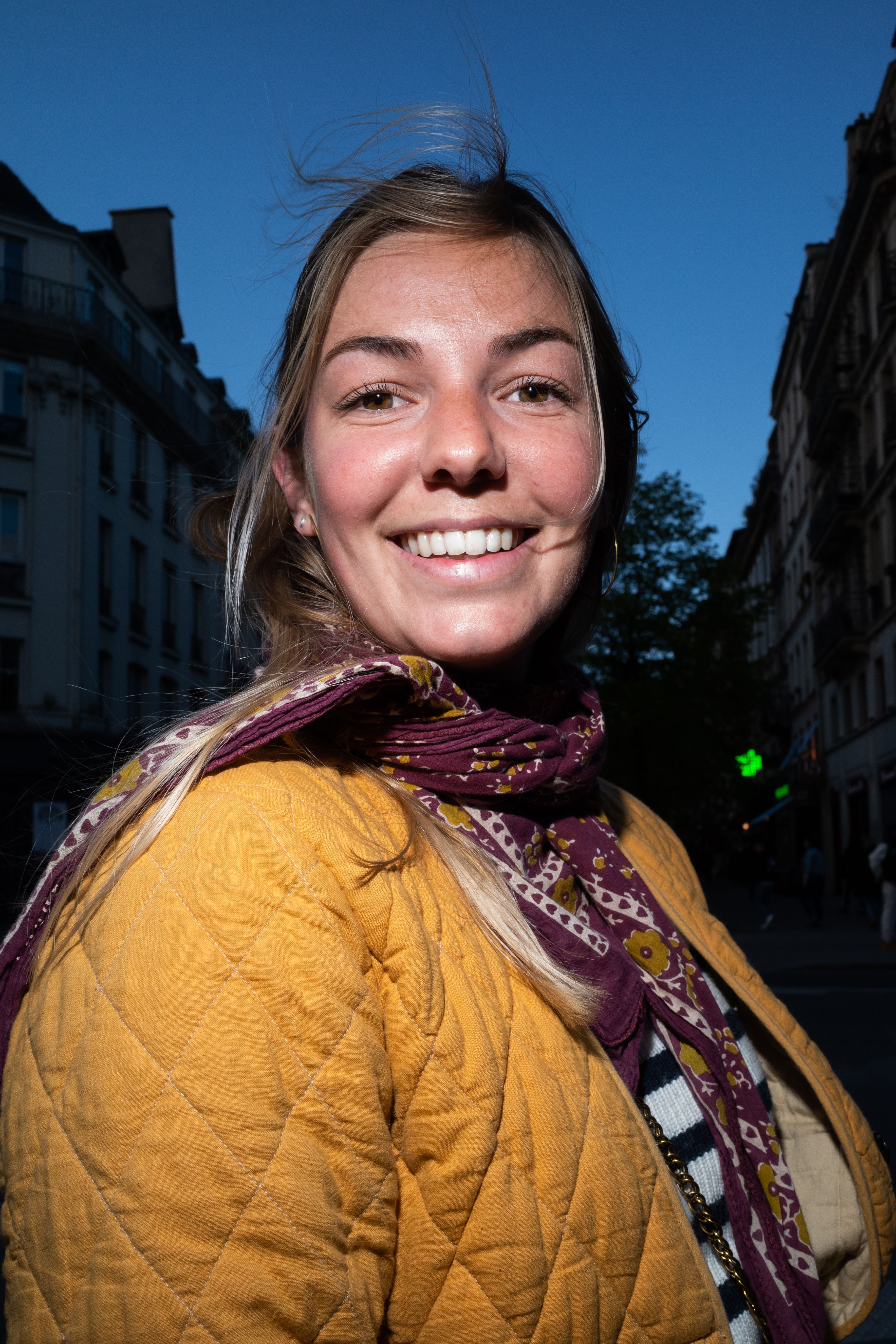 Smiling woman in autumn attire with a scarf in a vibrant city street setting.