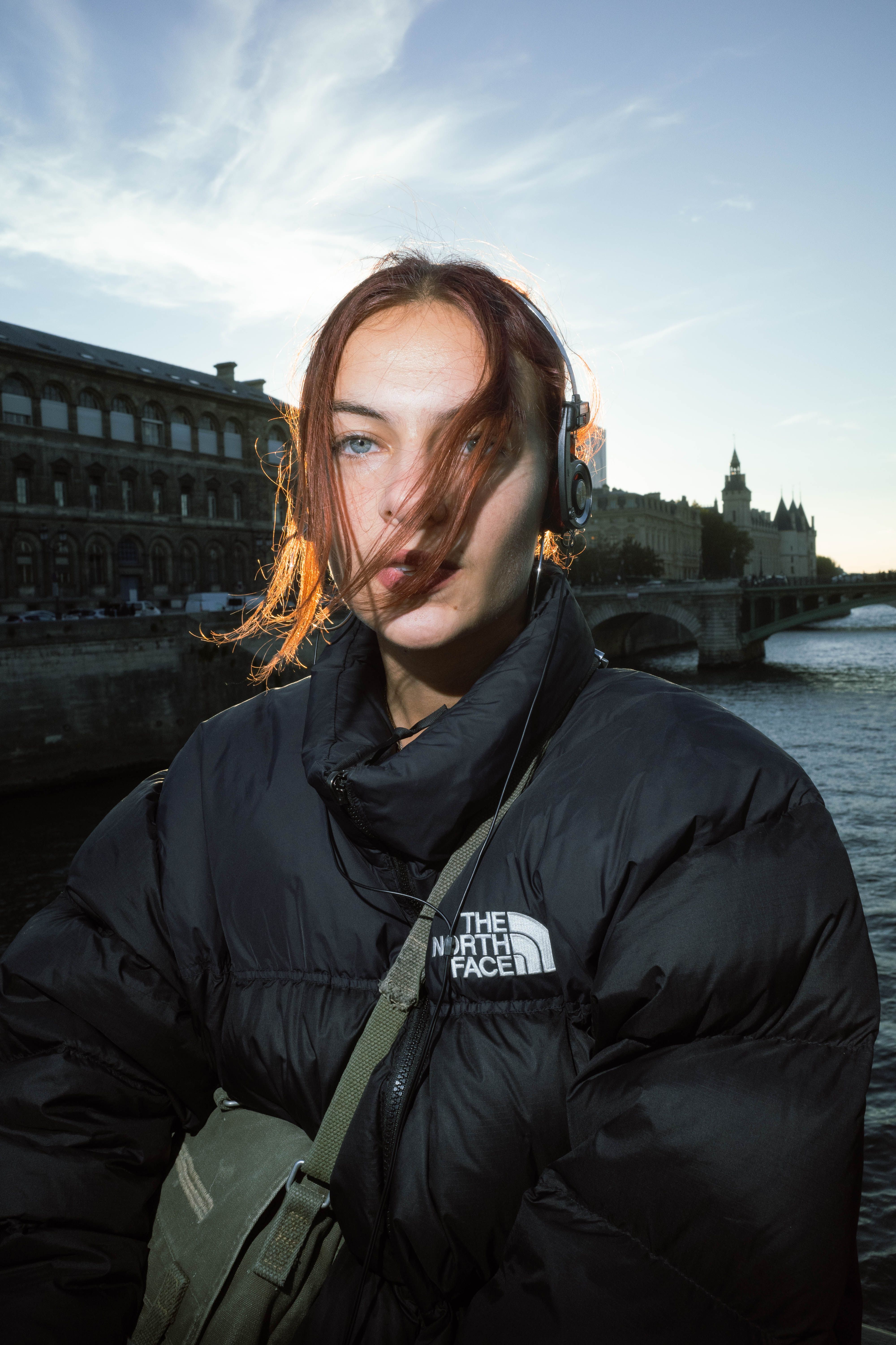Young woman in a jacket with headphones by a river during sunset.