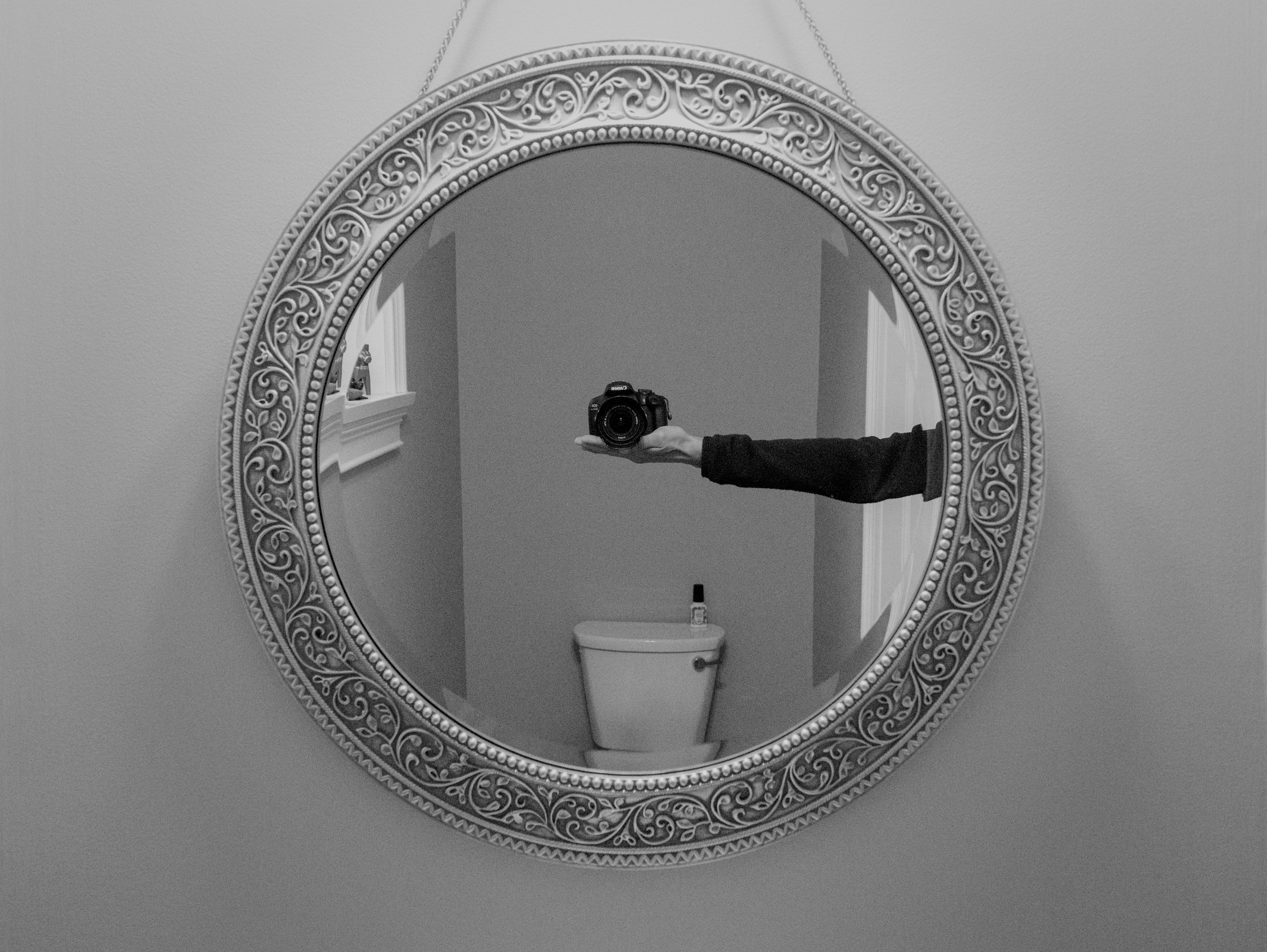 Intricate round mirror capturing a camera reflection in a monochrome bathroom setting.