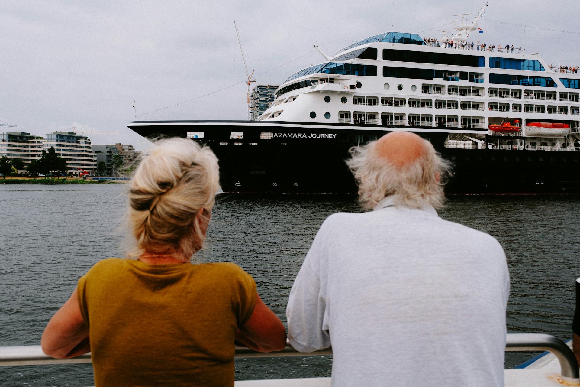 An elderly couple watches a large cruise ship in a bustling harbor.