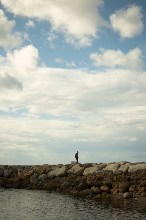 Free A person stands alone on rocky seaside under a dramatic cloudy sky, evoking solitude and peace. Stock Photo