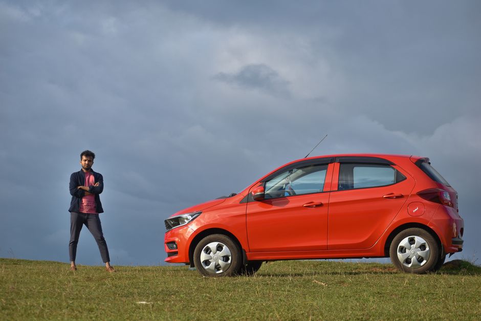 A man stands confidently beside a red car under a cloudy sky in Wayanad, India.