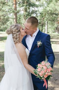 Bride and groom embracing and kissing in a sunny outdoor forest setting.