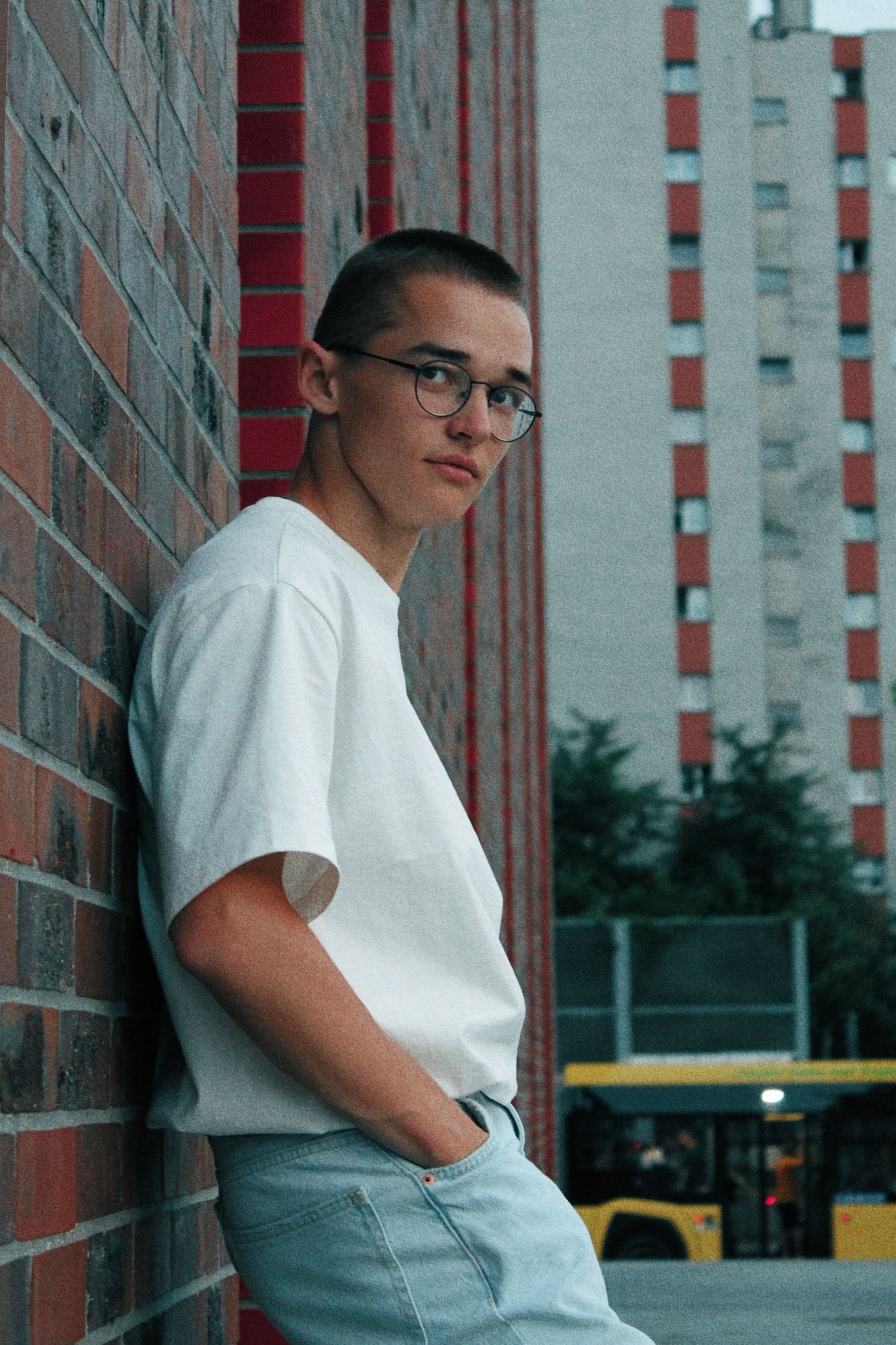 Teenage boy in eyeglasses stands by a brick wall in an urban setting in Katowice.