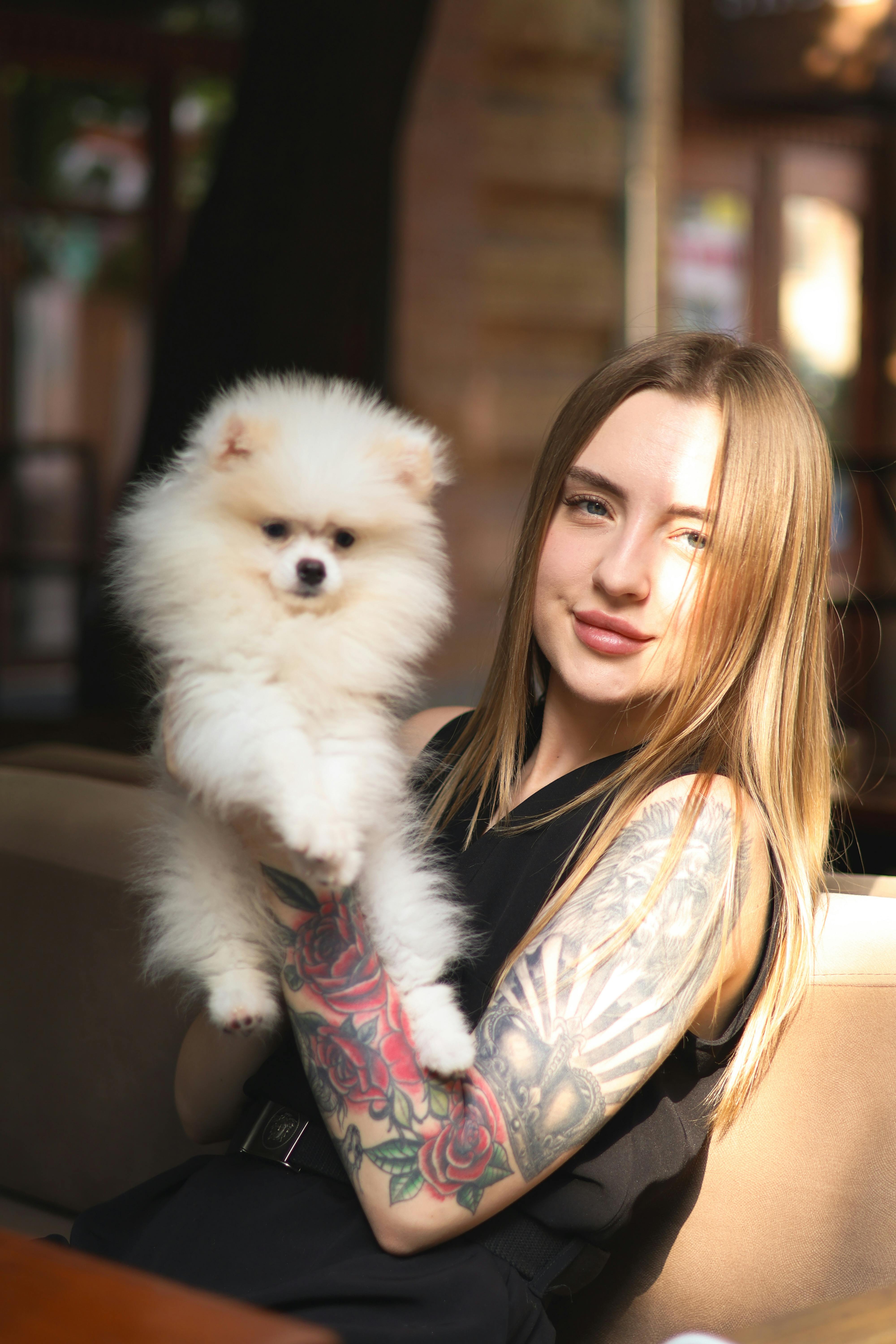 Young woman with tattoo holding a fluffy Pomeranian indoors, smiling warmly.