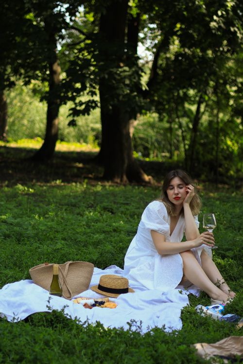 Free A woman in a white dress relaxes with wine at a picnic in a lush green park. Stock Photo