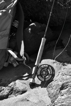 A black and white shot of fishing gear, including a rod and backpack, on rocky terrain by the coast.