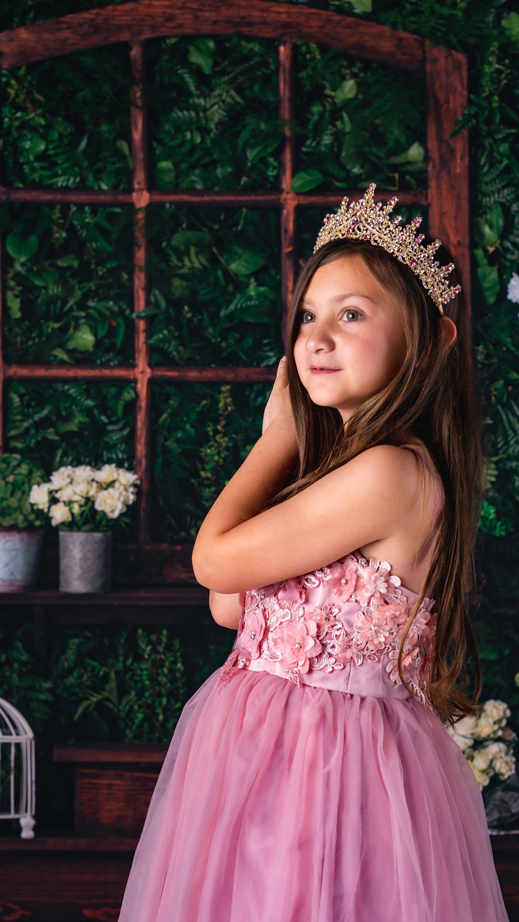 Adorable girl dressed as a princess in a pink gown with a crown, standing indoors.