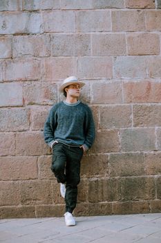 A fashionable man poses against a stone wall in Cusco, Peru, wearing a hat and casual outfit.